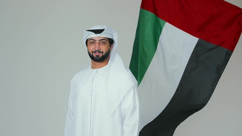 تعابير السعادة والسرور في المناسبات الوطنية، الأعياد و المناسبات الوطنية,  الاحتفال باليوم الوطني الإماراتي, مفهوم الحب و الانتماء للبلاد, بورتريه لرجل عربي خليجي إماراتي يرتدي الكندورة والغترة البيضاء يقوم بحمل علم الإمارات العربية المتحدة, خلفية بيضاء