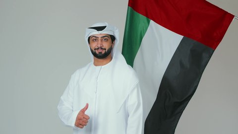 رفع اصبع الابهام بإيماءات الاعجاب بالمناسبات الوطنية، الأعياد و المناسبات الوطنية,  الاحتفال باليوم الوطني الإماراتي, مفهوم الحب و الانتماء للبلاد, بورتريه لرجل عربي خليجي إماراتي يرتدي الكندورة والغترة البيضاء يقوم بحمل علم الإمارات العربية المتحدة, خلفية بيضاء