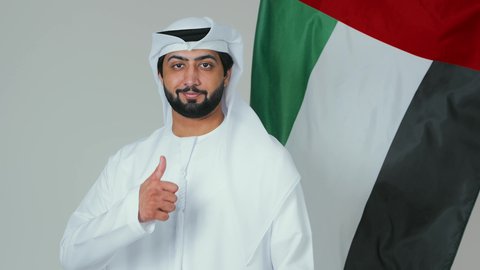 Raising the thumb with gestures of admiration for national occasions, holidays, and national events, celebrating the UAE National Day, the concept of love and belonging to the country, a portrait of an Arab Gulf Emirati man wearing the kandura and white ghutrah holding the flag of the United Arab Emirates, white background.