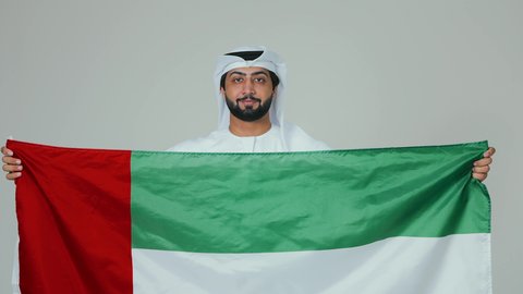 The concept of love and belonging to the country, happiness and joy in national occasions, celebrating the UAE National Day, holidays and national events, a portrait of an Arab Gulf Emirati man wearing the kandura and white ghutrah holding the flag of the United Arab Emirates, white background.
