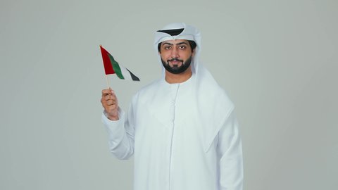 الأعياد و المناسبات الوطنية,  الاحتفال باليوم الوطني الإماراتي, مفهوم الحب و الانتماء للبلاد, السعادة والسرور في المناسبات الوطنية، بورتريه لرجل عربي خليجي إماراتي يرتدي الكندورة والغترة البيضاء يقوم بحمل علم الإمارات العربية المتحدة, خلفية بيضاء