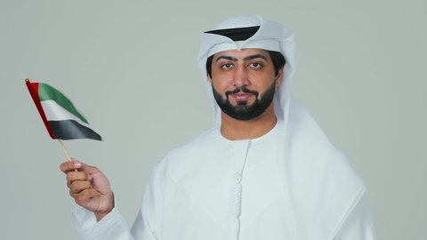 تعابير السعادة والسرور في المناسبات الوطنية، الأعياد و المناسبات الوطنية,  الاحتفال باليوم الوطني الإماراتي, مفهوم الحب و الانتماء للبلاد, بورتريه مقرب لرجل عربي خليجي إماراتي يرتدي الكندورة والغترة البيضاء يقوم بحمل علم الإمارات العربية المتحدة, خلفية بيضاء
