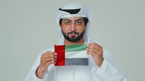 Expressions of happiness and joy during national occasions, holidays, and national events, celebrating the UAE National Day, the concept of love and belonging to the country, a close-up portrait of an Arab Gulf Emirati man wearing a kandura and a white ghutrah holding the flag of the United Arab Emirates, white background.