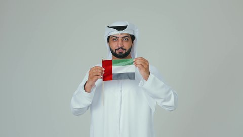 ايماءات السعادة والسرور في المناسبات الوطنية، الأعياد و المناسبات الوطنية,  الاحتفال باليوم الوطني الإماراتي, مفهوم الحب و الانتماء للبلاد, بورتريه لرجل عربي خليجي إماراتي يرتدي الكندورة والغترة البيضاء يقوم بحمل علم الإمارات العربية المتحدة, خلفية بيضاء