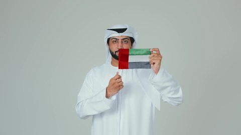 Holidays and national occasions, celebrating the UAE National Day, the concept of love and belonging to the country, happiness and joy in national occasions, a portrait of an Arab Gulf Emirati man wearing a kandura and a white ghutrah holding the flag of the United Arab Emirates, white background.