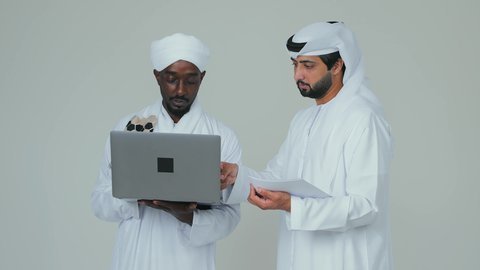 التعاون  بين الاصدقاء ،جهاز الحاسوب المحمول المتطور و الحديث،بناء علاقات اجتماعية ، بورتريه لرجل عربي خليجي اماراتي يرتدي الكندورة والغترة  يقف مع صديق سوداني يرتدي الجلابية و العمامة والشال يقومان بالعمل عبر اللاابتوب  ، خلفية بيضاء