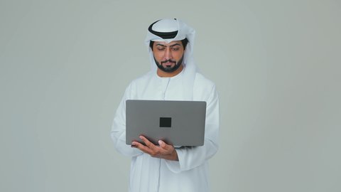 Using a modern and advanced technical device, completing work tasks remotely via a laptop, a portrait of an Emirati Gulf Arab man wearing a kandura and ghutrah holding a laptop in his hand, attention to external appearance, white background.