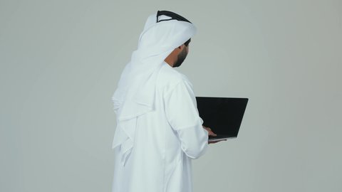 جهاز تقني حديث و متطور، انجاز مهام العمل عن بعد عبر جهاز اللابتوب ، بورتريه من الجانب  لرجل عربي خليجي اماراتي يرتدي الكندورة و الغترة  يحمل بيده الحاسوب المحمول، الاهتمام بالمظهر الخارجي، خلفية بيضاء