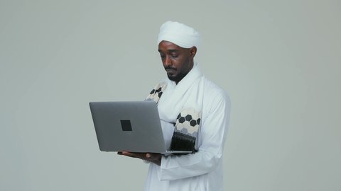 انجاز مهام العمل عن بعد عبر جهاز اللابتوب ،جهاز تقني حديث و متطور، بورتريه لرجل عربي سوداني يرتدي الجلابية ويضع العمامة والشال يحمل بيده الحاسوب المحمول، الاهتمام بالمظهر الخارجي، خلفية بيضاء