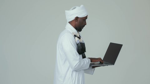 A modern and advanced technical device, accomplishing remote work tasks via a laptop, a side portrait of a Sudanese Arab man wearing a jalabiya and a turban, holding a laptop in his hand, with attention to external appearance, white background.
