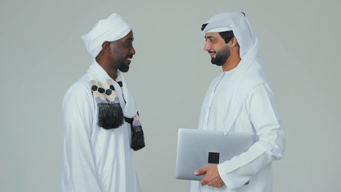 Chat and conversations between friends, advanced and modern laptop, building social relationships, a portrait of an Emirati Gulf Arab man wearing a kandura and ghutrah standing with a Sudanese friend wearing a jalabiya, turban, and shawl, white background.