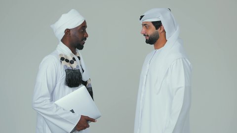 دردشة ومحادثات بين الاصدقاء ،جهاز الحاسوب المحمول المتطور و الحديث،بناء علاقات اجتماعية ، بورتريه لرجل عربي خليجي اماراتي يرتدي الكندورة والغترة  يقف مع صديق سوداني يرتدي الجلابية و العمامة والشال ، خلفية بيضاء