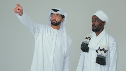 Pointing with the index finger at something specific, a portrait of an Emirati Gulf Arab man wearing a kandura and ghutrah standing with a Sudanese friend and pointing at something, standing confidently and paying attention to appearance, white background.