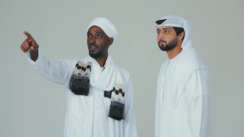 الاشارة باصبع السبابة الى شيء معين، بورتريه لرجل عربي سوداني يرتدي الجلابية ويضع العمامة والشال يقف مع صديق اماراتي  ويشير إلى شيء ما، الوقوف بثقة والاهتمام بالمظهر الخارجي، خلفية بيضاء