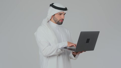 مهارة الايدي السريعة على لوحة المفاتيح، استخدام التقنيات الحديثة المتطورة، إدارة شؤون الأعمال وتنظيمها عن بعد، دمج التكنولوجيا في الحياة العملية، بورتريه جانبي لرجل عربي خليجي اماراتي يرتدي الكندورة والغترة يقوم بالعمل على الحاسب المحمول, خلفية بيضاء