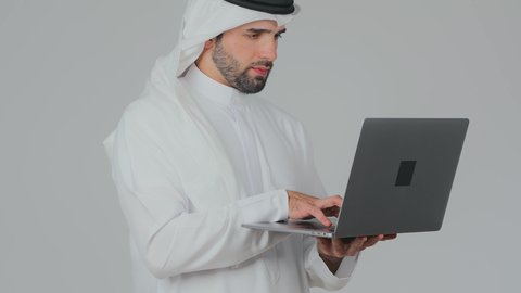 الايدي السريعة على لوحة المفاتيح، استخدام التقنيات الحديثة المتطورة، إدارة شؤون الأعمال وتنظيمها عن بعد، دمج التكنولوجيا في الحياة العملية، بورتريه جانبي لرجل عربي خليجي اماراتي يرتدي الكندورة والغترة يقوم بالعمل على الحاسب المحمول, خلفية بيضاء