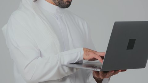 Working with advanced modern technologies, skillful hands on the keyboard, managing and organizing business affairs remotely, integrating technology into practical life, a close-up side portrait of an Emirati Gulf Arab man wearing a kandura and ghutrah working on a laptop, white background.
