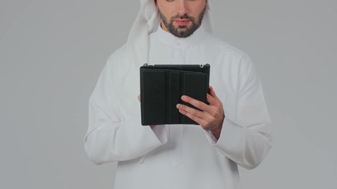 تطبيقات وبرامج الأجهزة اللوحية، استخدام التكنولوجيا والأجهزة الحديثة والمتطورة، دمج التقنية في الحياة اليومية، بورتريه مقرب لرجل عربي خليجي إماراتي يرتدي الكندورة والغترة يقف ويقوم بالنقر على جهاز التابلت، تصفح مواقع التواصل الاجتماعي عبر الانترنت، خلفية بيضاء