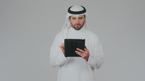 تصفح مواقع التواصل الاجتماعي عبر الانترنت، تطبيقات وبرامج الأجهزة اللوحية، استخدام التكنولوجيا والأجهزة الحديثة والمتطورة، دمج التقنية في الحياة اليومية، بورتريه لرجل عربي خليجي إماراتي يرتدي الكندورة والغترة يقف ويقوم بالنقر على جهاز التابلت، خلفية بيضاء