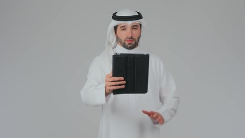 Video call via a tablet, browsing social media websites online, using modern and advanced technology and devices, integrating technology into daily life, a portrait of an Emirati Gulf Arab man wearing a kandura and ghutrah standing and tapping on a tablet device, white background.