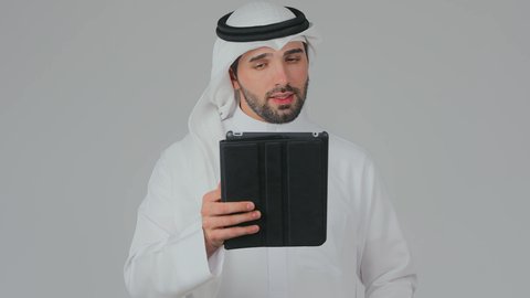 اجراء مكالمة فيديو عبر الجهاز اللوحي، تصفح مواقع التواصل الاجتماعي عبر الانترنت، استخدام التكنولوجيا والأجهزة الحديثة والمتطورة، دمج التقنية في الحياة اليومية، بورتريه لرجل عربي خليجي إماراتي يرتدي الكندورة والغترة يقف ويقوم بالنقر على جهاز التابلت، خلفية بيضاء