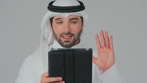 Greeting through a video call via a tablet, browsing social media, using modern and advanced technology and devices, integrating technology into daily life, a close-up portrait of an Emirati Gulf Arab man wearing a kandura and ghutrah standing and holding a tablet, white background.