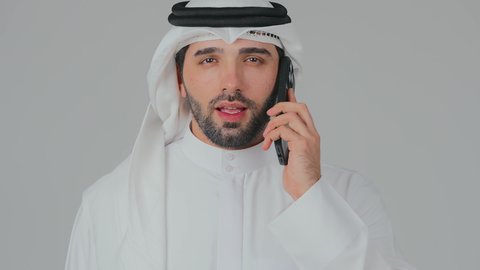 A phone call via a mobile device, browsing social media, using modern and advanced technology and devices, integrating technology into daily life, a close-up portrait of an Emirati Gulf Arab man wearing a kandura and ghutrah standing and holding a mobile phone, white background.