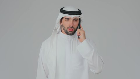 تعابير التعب والعتب خلال مكالمة هاتفية عبر الجهاز المحمول، تصفح مواقع التواصل الاجتماعي، استخدام التكنولوجيا والأجهزة الحديثة والمتطورة، دمج التقنية في الحياة اليومية، بورتريه مقرب لرجل عربي خليجي إماراتي يرتدي الكندورة والغترة يقف ويحمل الهاتف المحمول، خلفية بيضاء