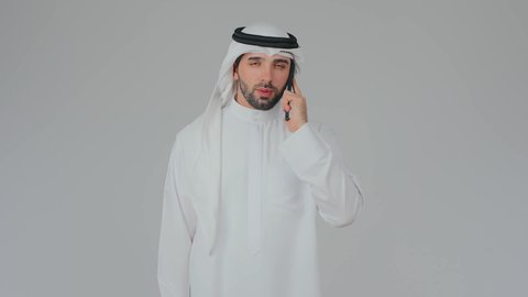المناقشة والتفاهم خلال مكالمة هاتفية عبر الجهاز المحمول، تصفح مواقع التواصل الاجتماعي، استخدام التكنولوجيا والأجهزة الحديثة والمتطورة، دمج التقنية في الحياة اليومية، بورتريه مقرب لرجل عربي خليجي إماراتي يرتدي الكندورة والغترة يقف ويحمل الهاتف المحمول، خلفية بيضاء