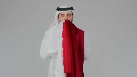 Loyalty and national belonging to the State of Qatar, commemorating the founding of the state, celebrating Qatar National Day on December 18, national cultures and symbols, love for the homeland and belonging to it, a portrait of an Arab Gulf Qatari man wearing traditional attire and a ghutrah holding the Qatari flag and smelling it with pride and honor, white background.