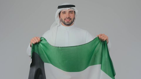 اليوم الوطني الاماراتي 2 ديسمبر، الاحتفال باليوم الوطني الإماراتي, الأعياد و المناسبات الوطنية,  مفهوم الحب و الانتماء للبلاد, بورتريه مقرب لرجل عربي خليجي إماراتي يرتدي الكندورة والغترة البيضاء يقوم بحمل علم الإمارات العربية المتحدة, خلفية بيضاء