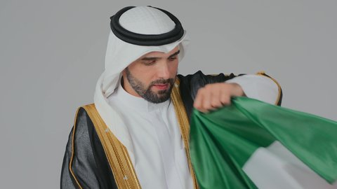 ارتداء الازياء الشعبية في المناسبات الوطنية،  الاحتفال باليوم الوطني الإماراتي, الأعياد و المناسبات الوطنية, بورتريه مقرب لرجل عربي خليجي إماراتي يرتدي الكندورة والغترة والبشت يقوم بوضع علم الإمارات العربية المتحدة على الاكتاف, خلفية بيضاء
