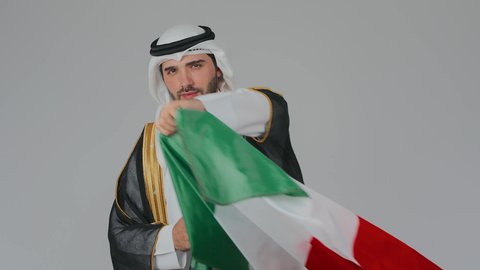 النظر الى الكاميرا بايماءات الاعتزاز والفخر, الاحتفال باليوم الوطني الإماراتي, الأعياد و المناسبات الوطنية, بورتريه مقرب لرجل عربي خليجي إماراتي يرتدي الكندورة والغترة والبشت البيضاء يقوم بوضع علم الإمارات العربية المتحدة على الاكتاف, خلفية بيضاء