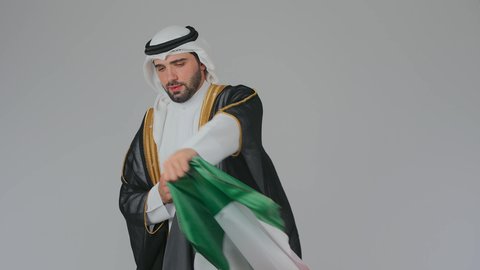 Love of the homeland and pride in it, celebrating the UAE National Day, national holidays and occasions, the concept of love and belonging to the country, a close-up portrait of an Emirati Gulf Arab man wearing the kandura, ghutrah, and bisht placing the flag of the United Arab Emirates on his shoulders, white background.