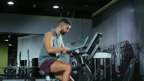 التدرب على جهاز العجلة الهوائية بتعابير الحماس, المحافظة على نظام الحياة الصحي،  مفهوم الرشاقة واللياقة البدنية، انشطة ورياضات لتقوية العضلات و الاعصاب، رجل عربي خليجي سعودي يرتدي الملابس الرياضية يتمرن في النادي الرياضي