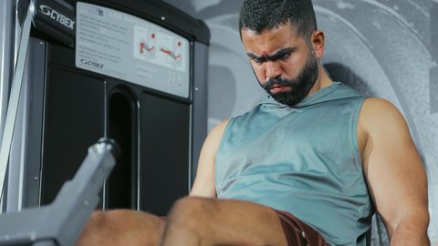 Muscle control over weight, attention to health and fitness, use of exercise equipment: a young Saudi Arabian man wearing sportswear exercises on a leg exercise machine at the gym; a healthy lifestyle; maintaining physical balance.