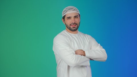 الوقوف باستقامة وايدي متشابكة ، مفهوم الرجولة والثقة بالنفس, بورتريه لرجل عربي خليجي إماراتي يرتدي الكندورة والقبعة البيضاء ينظر الى الكاميرا بتعابير السرور، الأناقة والاهتمام بالمظهر الخارجي، خلفية ملونة.