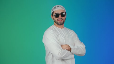 الأناقة والاهتمام بالمظهر الخارجي، الوقوف باستقامة وايدي متشابكة ، مفهوم الرجولة والثقة بالنفس, بورتريه لرجل عربي خليجي إماراتي يرتدي الكندورة والقبعة البيضاء والنظارة السوداء ينظر الى الكاميرا بتعابير السرور، خلفية ملونة.