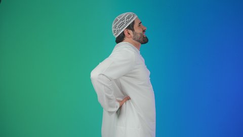 الإحساس بالألم والإصابة بمرض الديسك، آلام الظهر والعمود الفقري والتهاب المفاصل، بورتريه من الجانب لرجل عربي خليجي إماراتي يرتدي الكندورة الإماراتية والطاقيه يقف بصعوبة ويمسك ظهره بيديه بإيماءات المعاناة،  خلفية ملونه