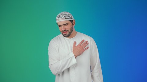 الاحساس بذبحه صدريه، التعب والإرهاق والضغوطات النفسية, الشعور في زيادة معدل ضربات القلب,بورتريه مقرب لرجل عربي خليجي اماراتي يرتدي الكندورة والقبعة البيضاء يضع يده على قلبه ويشعر بحالة سيئة من الوخز والألم, اضطرابات وأمراض قلبية, خلفية ملونة