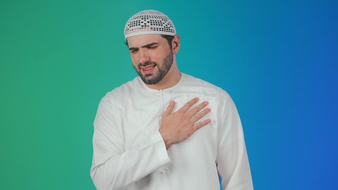 الشعور في زيادة معدل ضربات القلب, التعب والإرهاق والضغوطات النفسية, الاحساس بذبحه صدريه، بورتريه مقرب لرجل عربي خليجي اماراتي يرتدي الكندورة والقبعة البيضاء يضع يده على قلبه ويشعر بحالة سيئة من الوخز والألم, اضطرابات وأمراض قلبية, خلفية ملونة