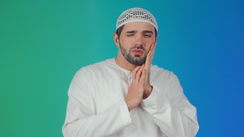 مفهوم تسوس الأضراس, الإنتفاخ والإلتهاب بالفم واللثة، بورتريه لشاب عربي خليجي  اماراتي يرتدي الكندورة والقبعة البيضاء يضع يده على وجهه بإيماءات الألم ووجع الأسنان، ضرورة الذهاب للطبيب والحصول على العلاج، خلفية ملونة