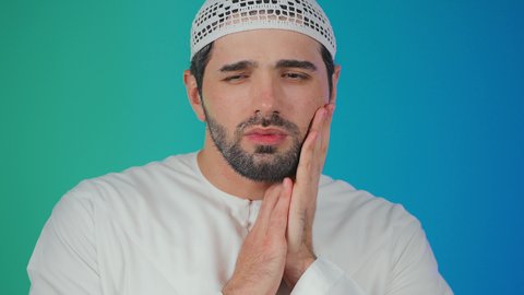 الإنتفاخ والإلتهاب بالفم واللثة، مفهوم تسوس الأضراس, بورتريه لشاب عربي خليجي  اماراتي يرتدي الكندورة والقبعة البيضاء يضع يده على وجهه بإيماءات الألم ووجع الأسنان، ضرورة الذهاب للطبيب والحصول على العلاج، خلفية ملونة
