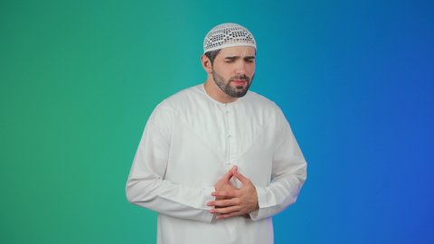الحاجة إلى العلاج، المعاناة من آلام البطن والمعدة، أمراض والتهابات الجهاز الهضمي،بورتريه لرجل عربي خليجي إماراتي يرتدي الكندورة والطاقيه يضع يديه على بطنه بتعابير التعب والإرهاق والإحساس بالمرض، خلفية ملونة