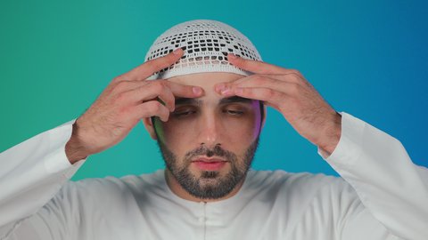 الاجهاد والضغط النفسي, الاحساس بالشقيقية والصداع النصفي, الشعور بالإرهاق وفقدان التركيز, بورتريه لرجل عربي خليجي اماراتي يرتدي  الكندورة والقبعة البيضاء يضع يديه على رأسه ويغمض عينيه بإيماءات ألم الرأس الشديد, خلفية ملونة