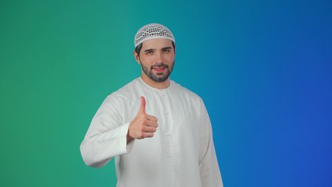 بورتريه مقرب لرجل عربي خليجي  اماراتي يرتدي الكندورة والطاقية يرفع يده للأعلى بإيماءات الرضا والقبول والأعجاب, النظر الى الكاميرا بتعابير السرور, التعبير عن الجودة والإتقان,خلفية ملونة.