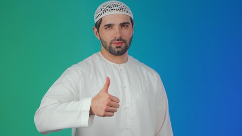 النظر الى الكاميرا بتعابير السرور, التعبير عن الجودة والإتقان, بورتريه مقرب لرجل عربي خليجي  اماراتي يرتدي الكندورة والطاقية يرفع يده للأعلى بإيماءات الرضا والقبول والأعجاب, خلفية ملونة.