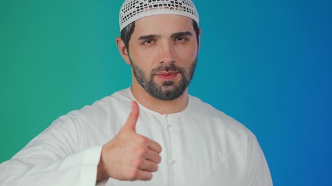 التعبير عن الجودة والإتقان, بورتريه مقرب لرجل عربي خليجي  اماراتي يرتدي الكندورة والطاقية يرفع يده للأعلى بإيماءات الرضا والقبول والأعجاب, النظر الى الكاميرا بتعابير السرور, خلفية ملونة.