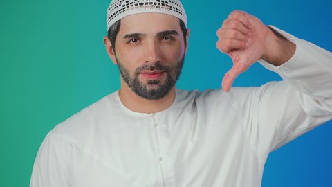 الإشارة بإصبع الإبهام للاسفل دليل الرفض، حركة يد وايماءات عدم الإعجاب والموافقة، بورتريه لرجل عربي اماراتي يرتدي الكندورة والطاقية يشير بايماءات عدم القبول، خلفية ملونة