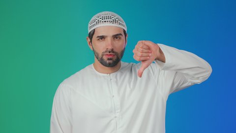 بورتريه لرجل عربي اماراتي يرتدي الكندورة والطاقية يشير بايماءات عدم القبول، الإشارة بإصبع الإبهام للاسفل دليل الرفض، حركة يد وايماءات عدم الإعجاب والموافقة،خلفية ملونة
